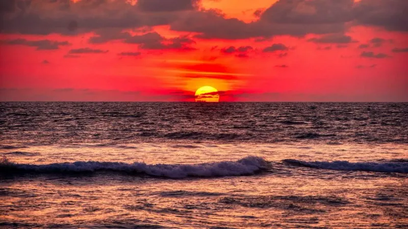 Langit senja berwarna merah menyala di atas laut lepas dengan ombak kecil menghampiri pantai